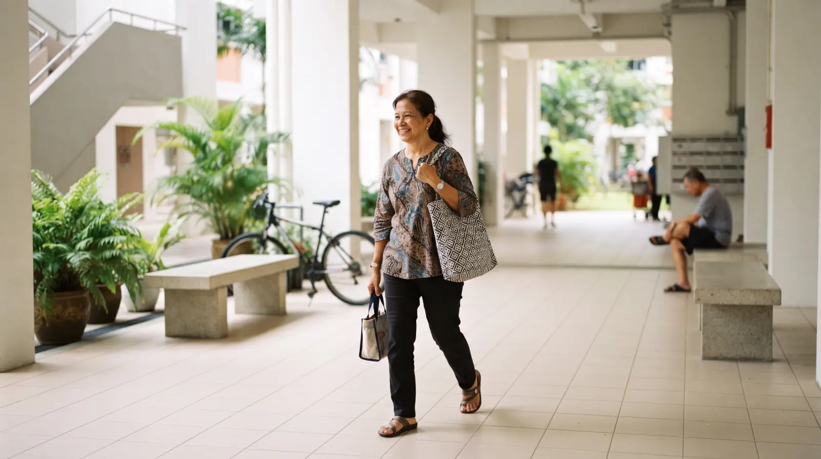 A woman in her fifties walking through a clean HDB void deck carrying a cloth shopping bag